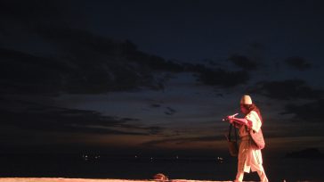 Indígena arhuaco, con un sombrero tradicional, toca una flauta al atardecer en Santa Marta. La figura se recorta contra el cielo oscuro y una tenue línea de horizonte.