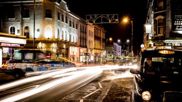 Calle en Sutton, Londres. Se aprecian edificios iluminados y las estelas de luz de vehículos en movimiento, incluyendo un taxi negro a la derecha.  