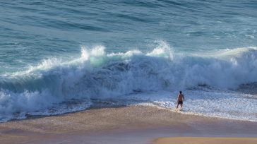 La imagen resalta el contraste entre la fragilidad humana y la inmensidad del mar, representado por una ola de gran tamaño rompiendo cerca de la orilla. &nbsp;