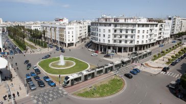 Una vista aérea de Rabat  muestra la  concurrida  Avenida Mohamed V, con una fuente central de agua en el medio. Un tranvía circula junto a la rotonda y muchos vehículos [&hellip;]
